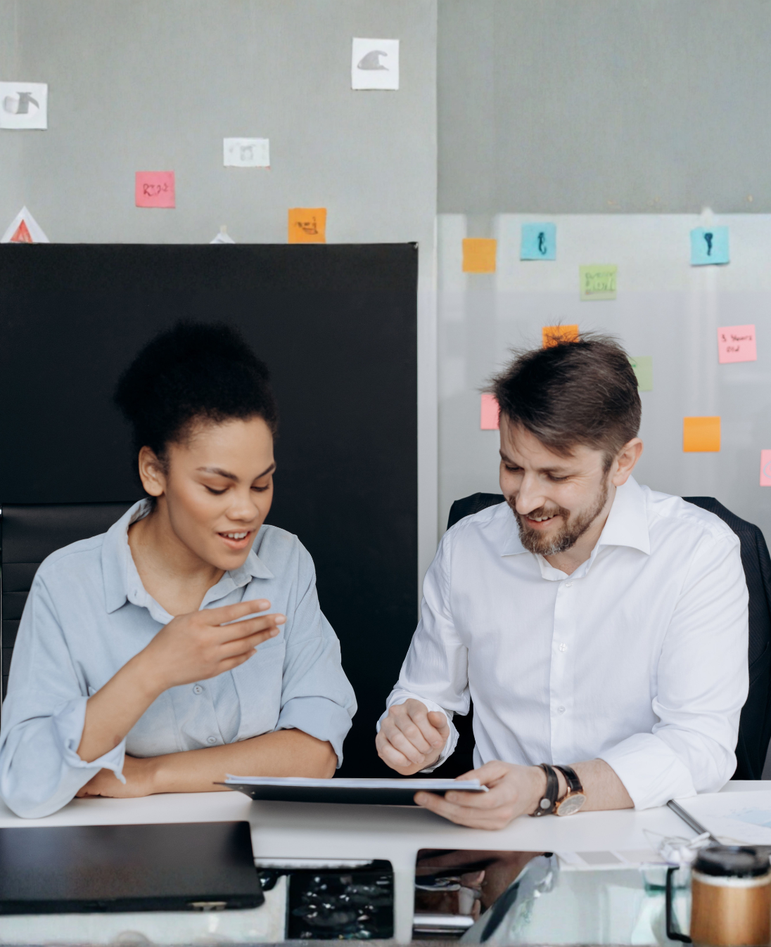 Two colleagues sitting side by side at a desk, looking down at a tablet the man is holding while the woman gestures as she speaks, in an office with sticky notes on the wall behind them.